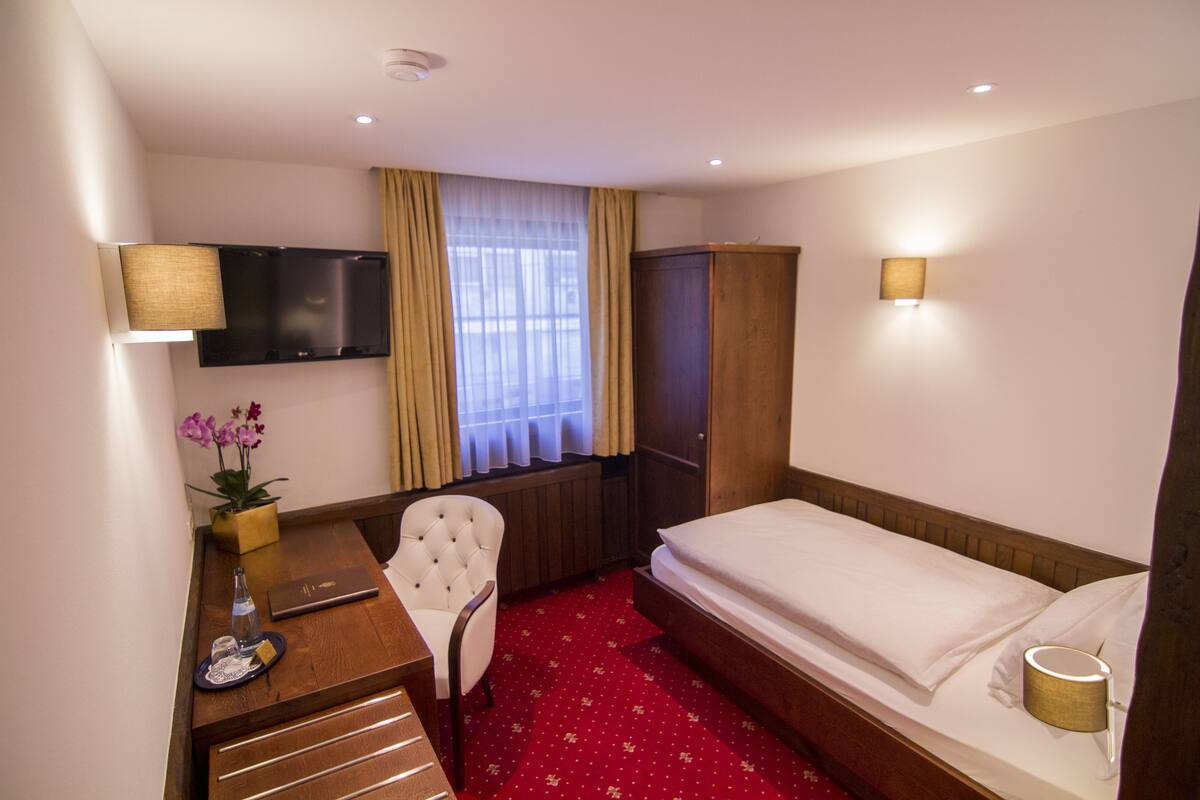 Habitación de hotel con cama, escritorio con silla, televisor montado en la pared, armario y ventana con cortinas.