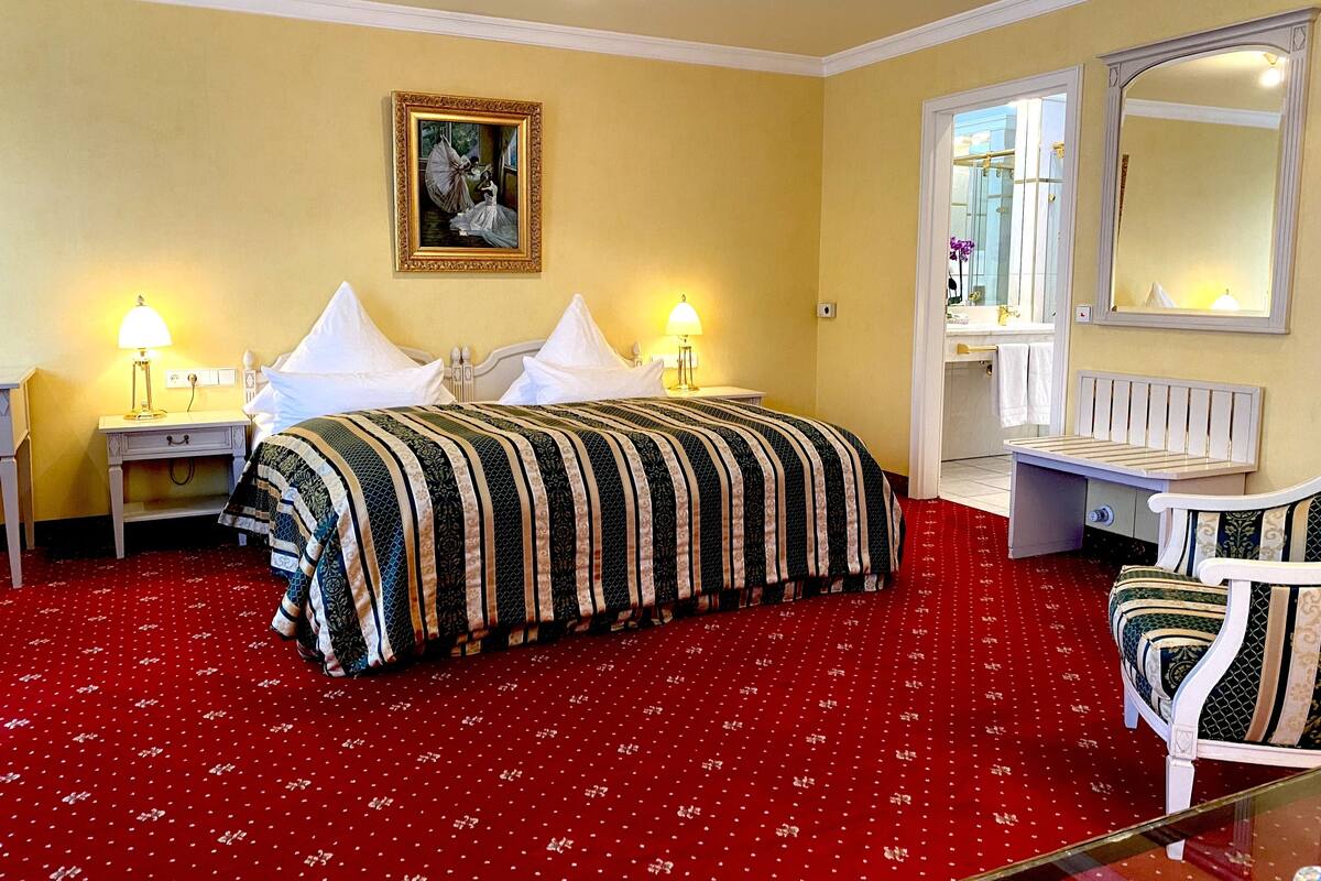 Habitación de hotel con una cama grande, dos mesitas de noche con lámparas, un espejo, un banco y suelo cubierto de alfombra roja.