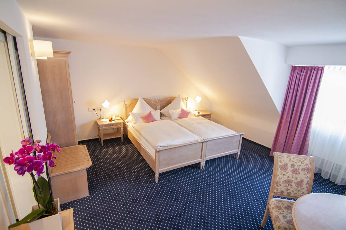 Una habitación de hotel con una cama, mesitas de noche, una silla y una pequeña mesa con una planta.