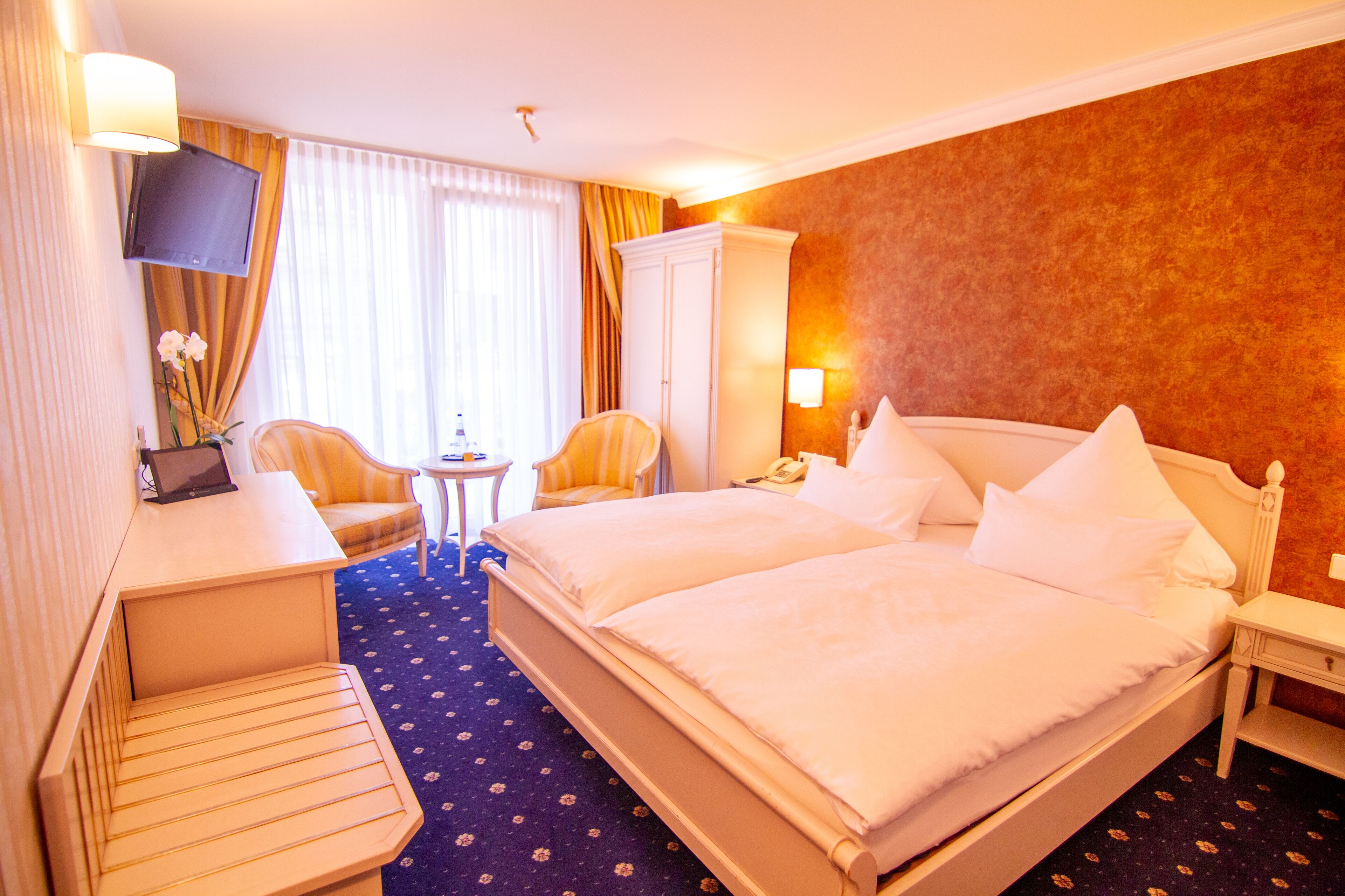 A hotel room with a double bed, a desk with a computer, a chair, a small table, and a TV mounted on the wall.