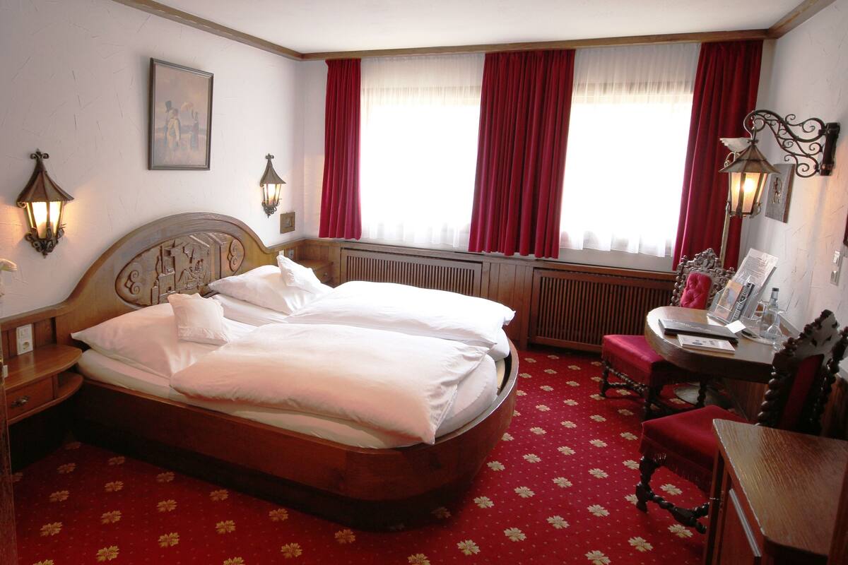 Habitación de hotel con cama doble, un escritorio con silla, un radiador y una ventana con cortinas rojas.