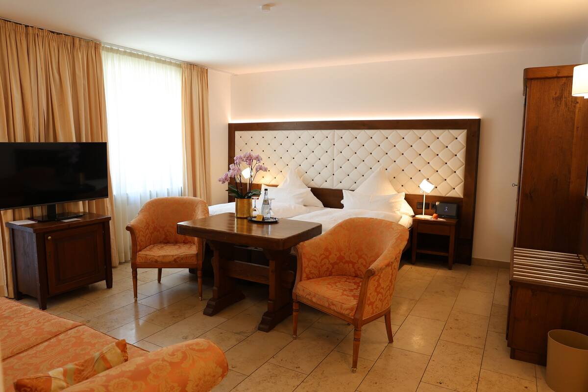 Una habitación de hotel con una cama grande, un televisor sobre un mueble, dos sillones, una mesita de noche de madera con lámparas y un armario de madera.