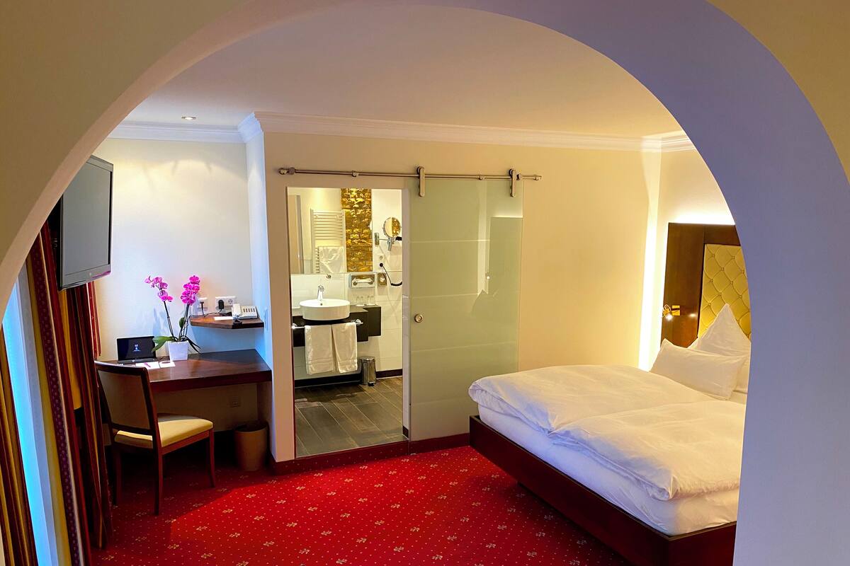 Habitación de hotel con cama, escritorio, silla, televisor y baño visible a través de un arco.