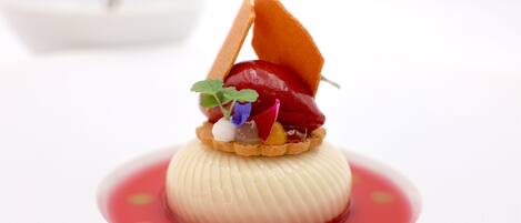 A dessert with a creamy base, topped with a fruit and a leaf, on a white plate with a red sauce.