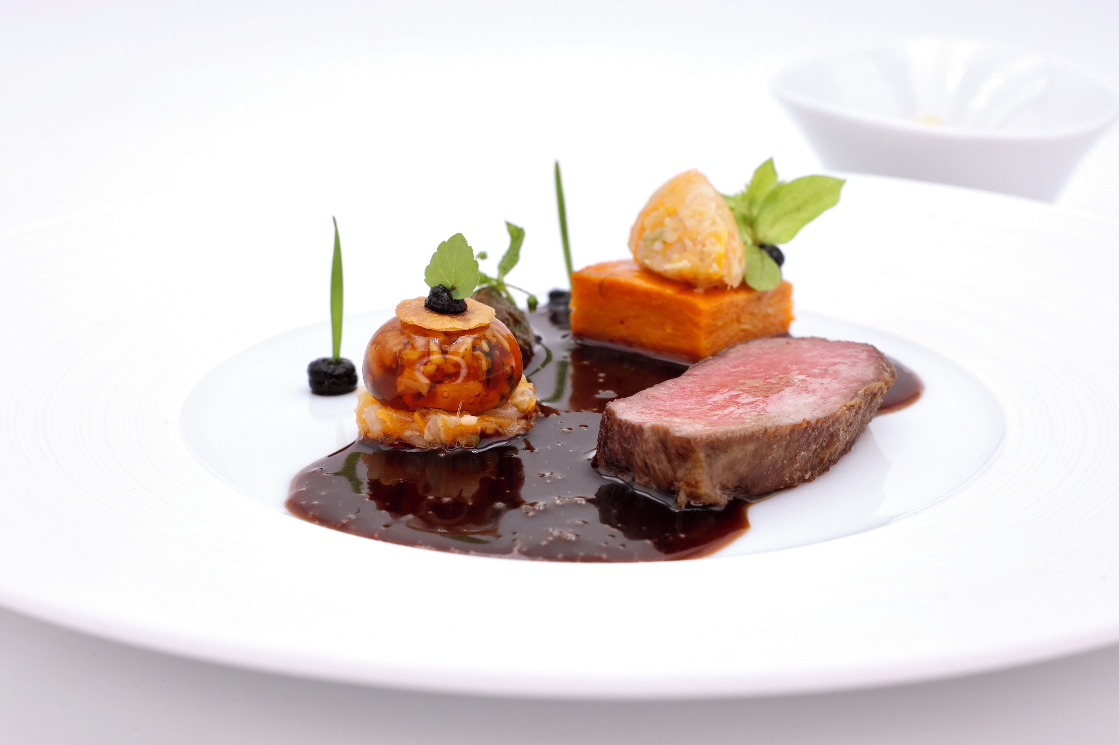 A white plate with a piece of steak, a small round dish with a garnish, and a square dish with a cube of food.