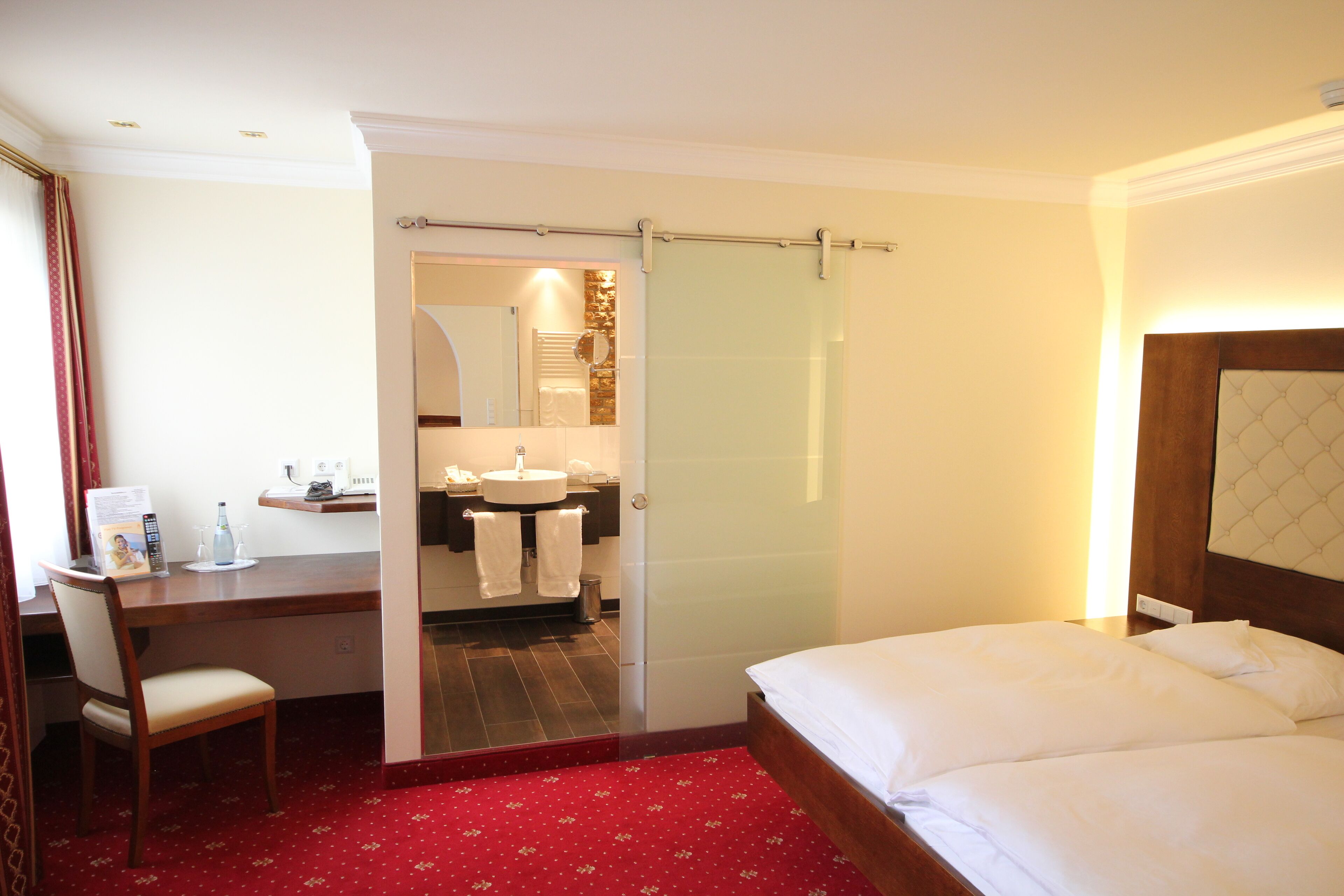 Habitación de hotel con cama, escritorio con silla, baño con lavamanos y espejo, y alfombra roja.