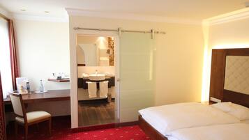 Habitación de hotel con cama, escritorio con silla, baño con lavamanos y espejo, y alfombra roja.