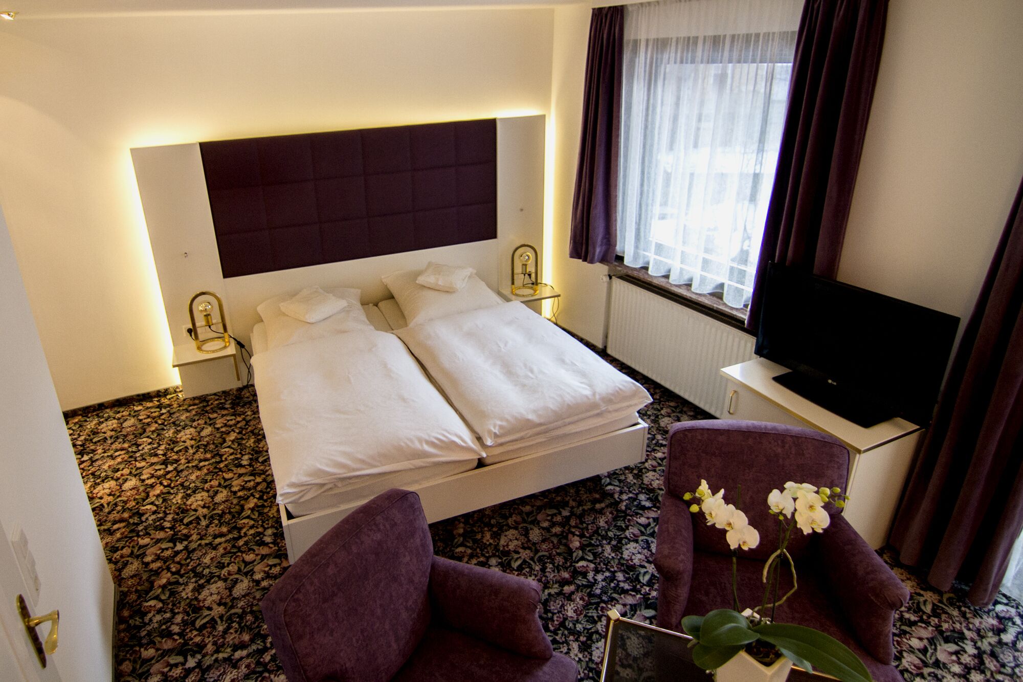 Ein Hotelzimmer mit einem Doppelbett, einem Fernseher, einem Sessel und einer Topforchidee.