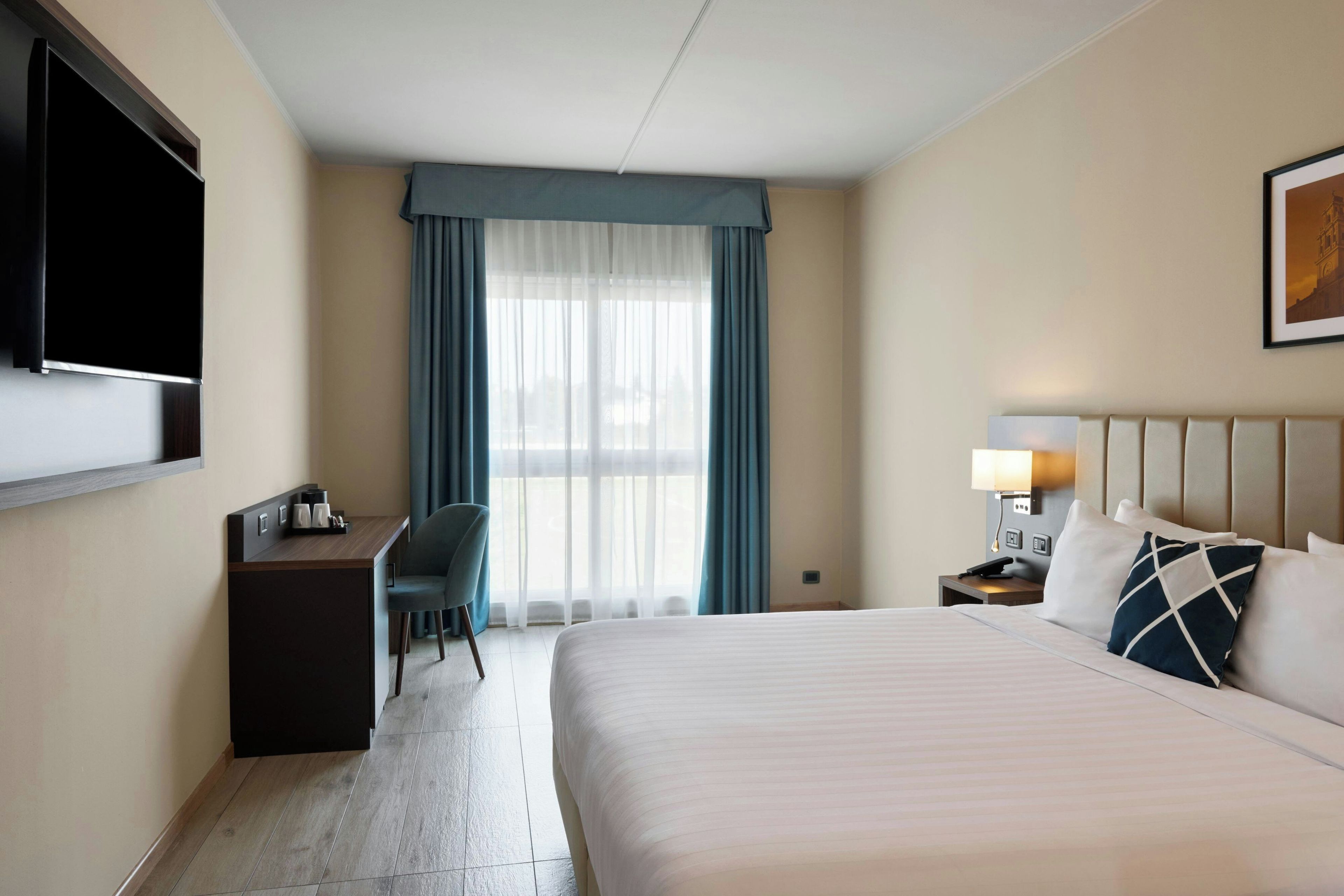 A hotel room with a bed, a desk, a chair, a TV, a window with curtains, and a wall-mounted lamp.