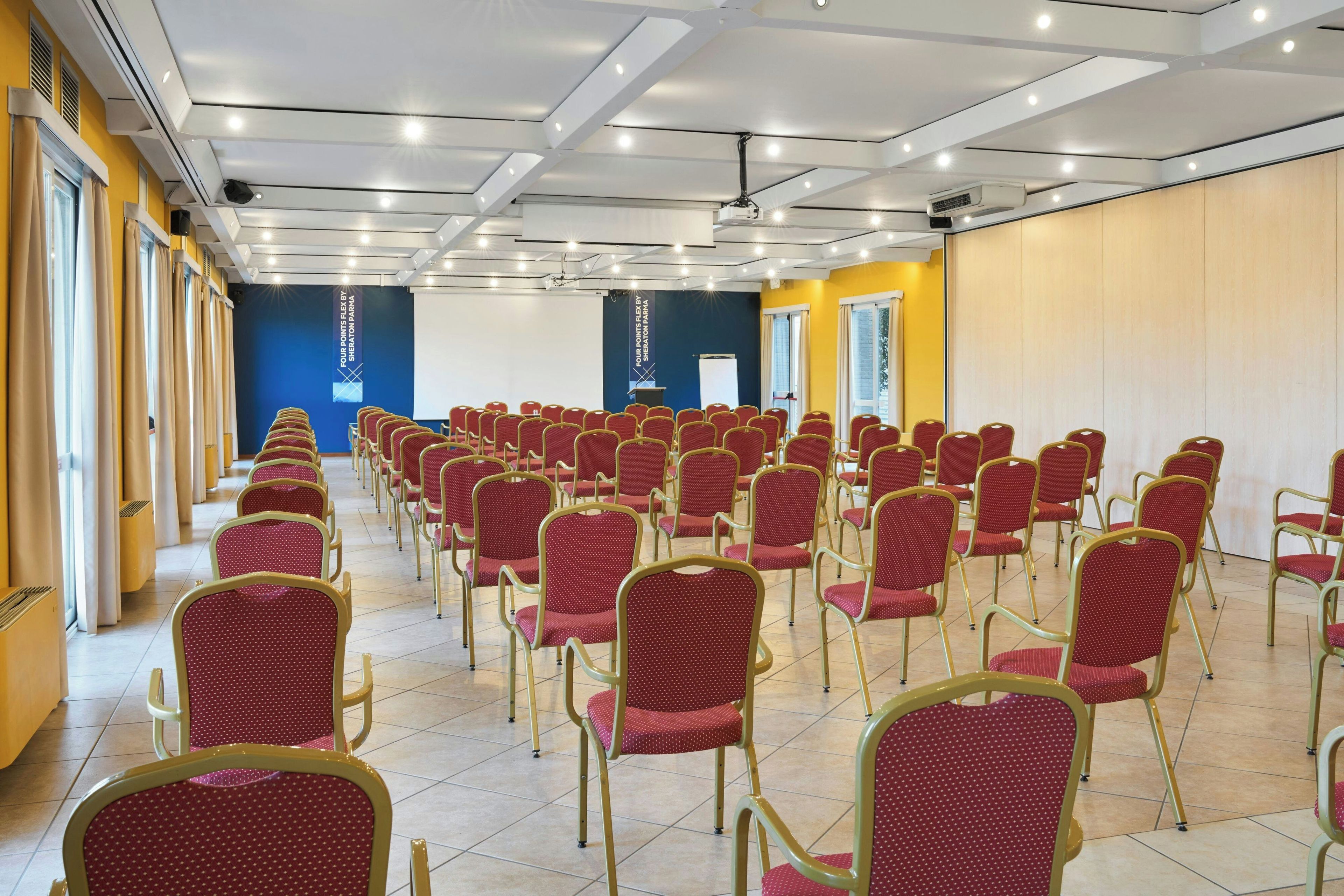 Una sala conferenze con file di sedie rosse, un podio bianco e una parete blu con uno schermo per proiettore.