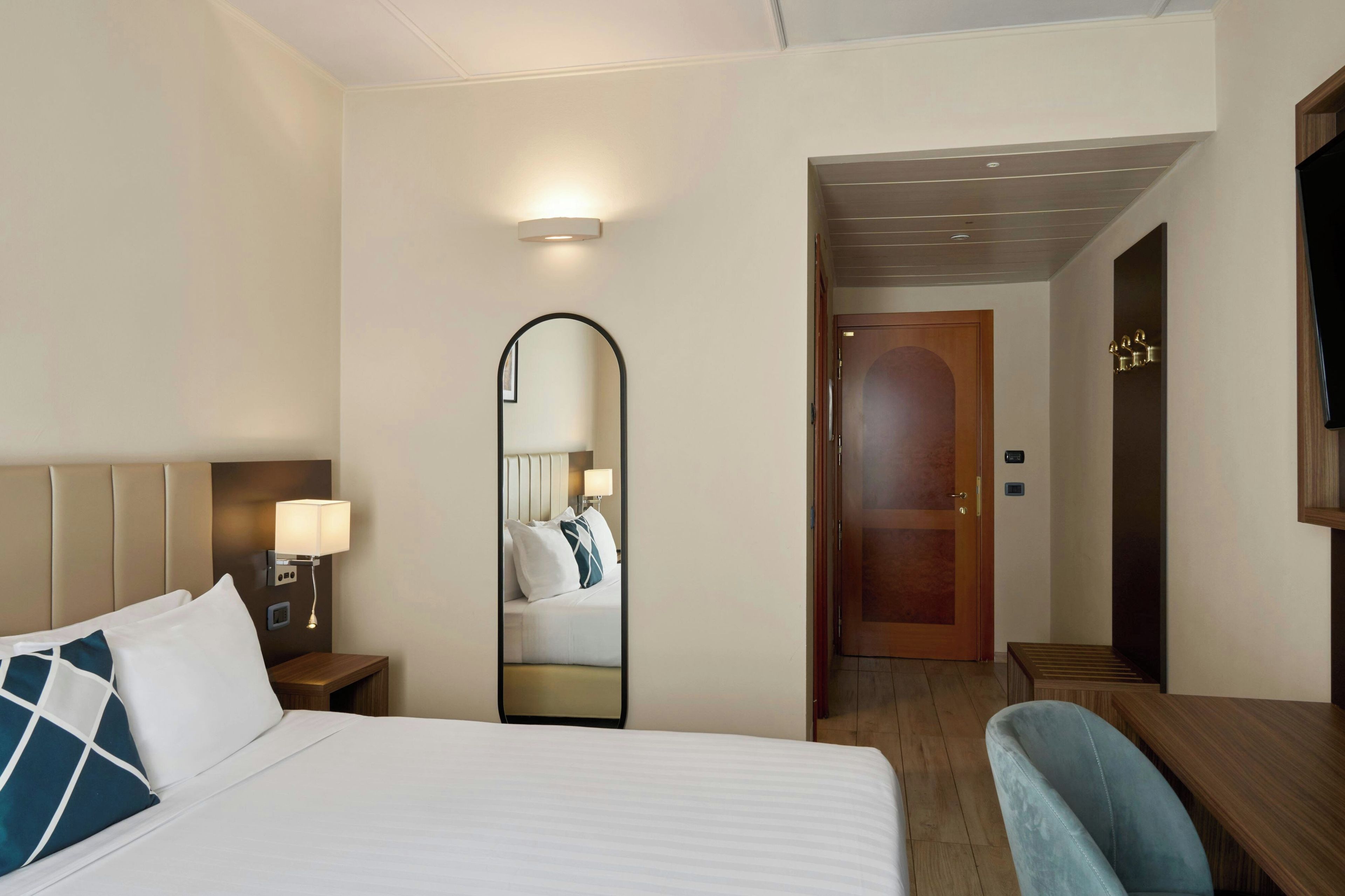 A hotel room with a bed, bedside table, lamp, mirror, and a wooden door leading to a hallway.