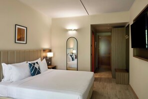 A hotel room with a large bed, a bedside table with a lamp, a mirror, a television, and a framed picture on the wall.