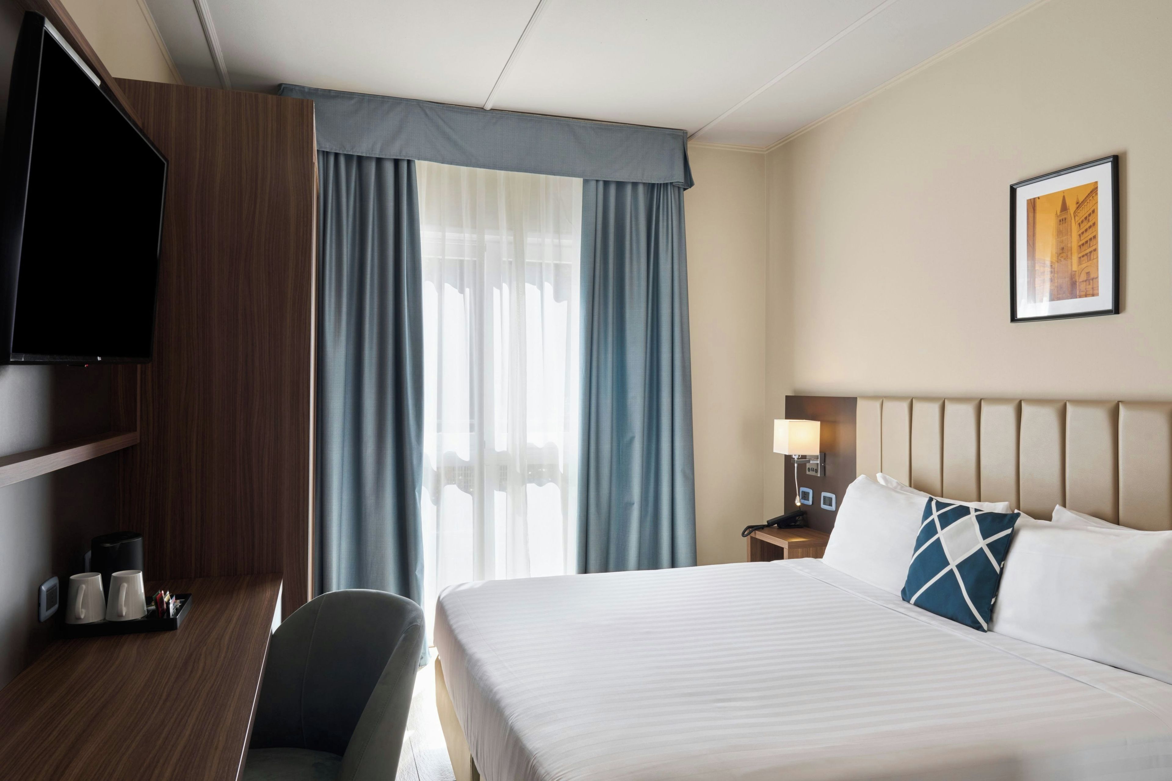 A hotel room with a bed, a desk, a chair, a television, a nightstand, a lamp, and a framed picture on the wall.