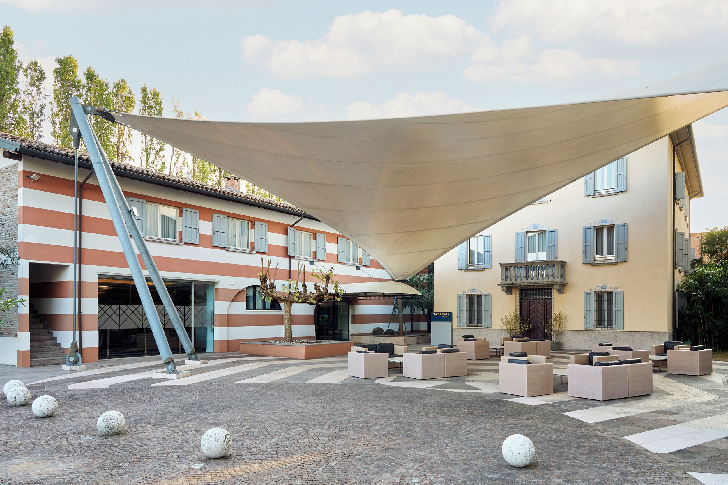 Une cour dotée d’un espace extérieur aménagé avec des sièges, d’une grande structure offrant de l’ombre, et d’un bâtiment aux murs rayés et aux volets bleus.