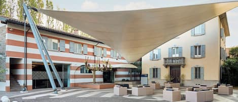 Un patio con mobiliario exterior, una amplia estructura de sombra y un edificio con paredes a rayas y contraventanas azules.