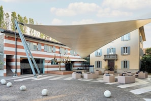 A courtyard with outdoor seating, a large shade structure, and a building with striped walls and blue shutters. - Four Points Flex by Sheraton Parma (Parma)