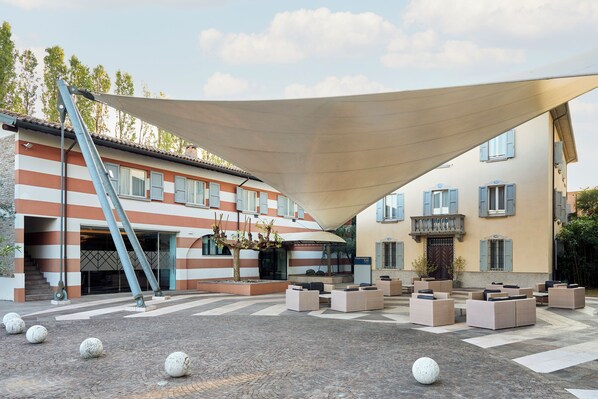 A courtyard with outdoor seating, a large shade structure, and a building with striped walls and blue shutters.