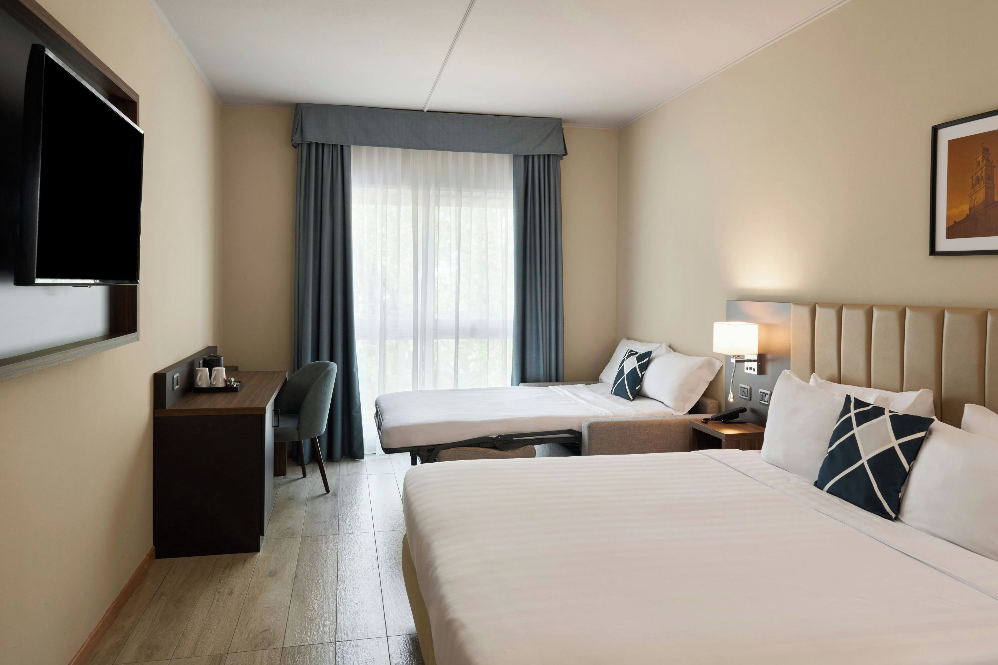 Una habitación de hotel con dos camas, un escritorio con silla, televisión y ventana con cortinas.
