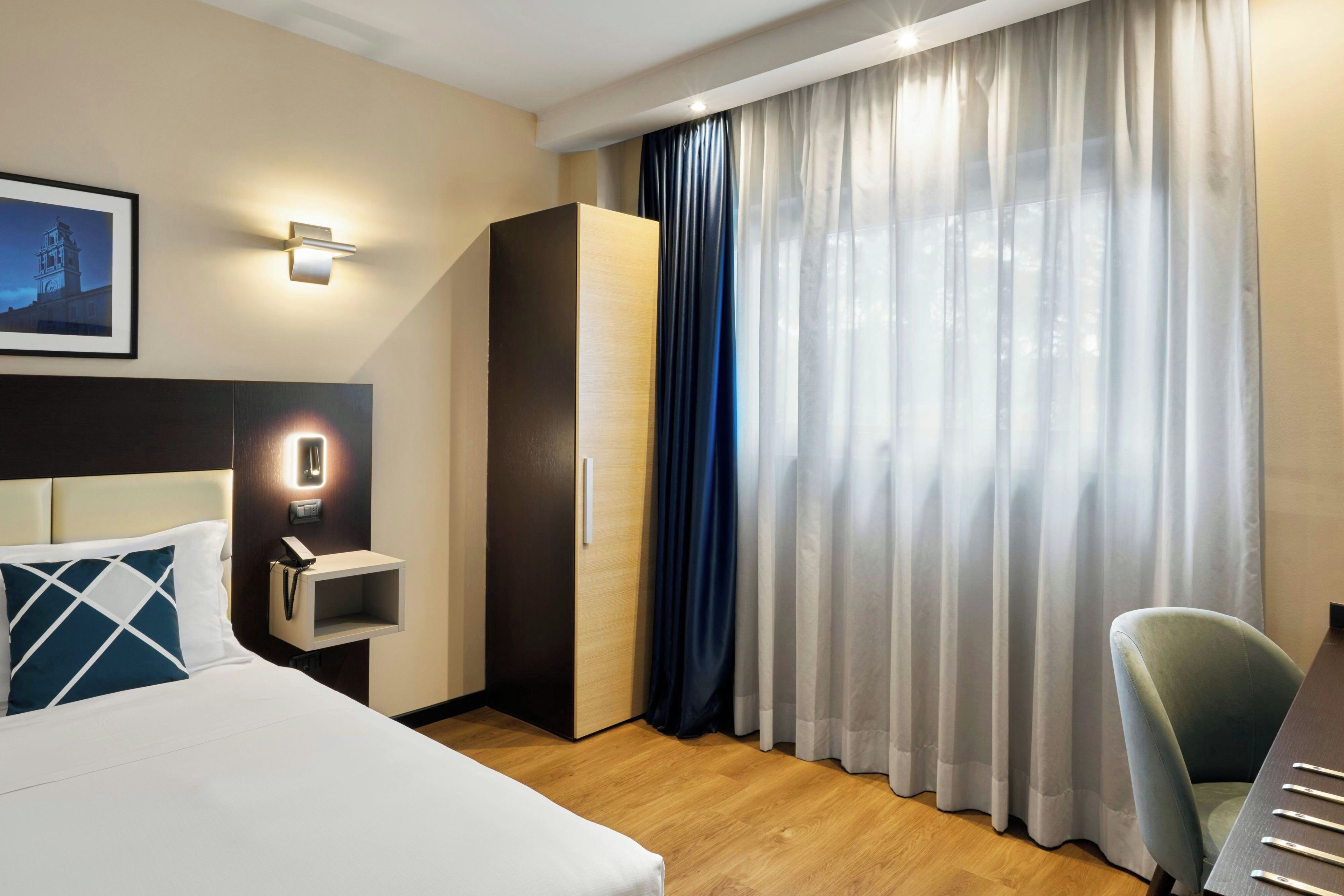 Een hotelkamer met een bed, een bureau, een kledingkast, een telefoon aan de muur en een raam met gordijnen.