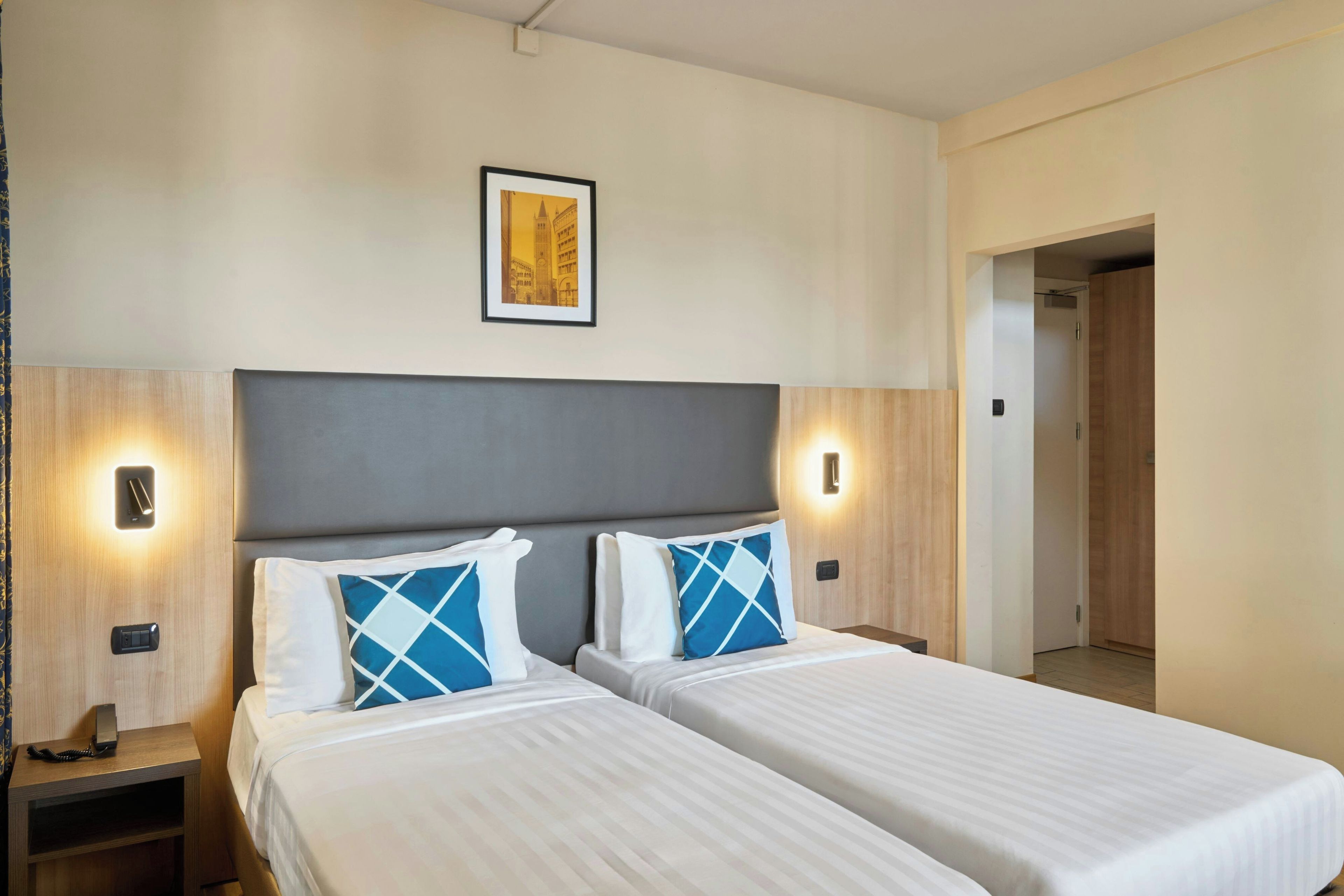 A hotel room with two beds, a headboard, bedside lamps, a framed picture on the wall, and a door leading to another room.