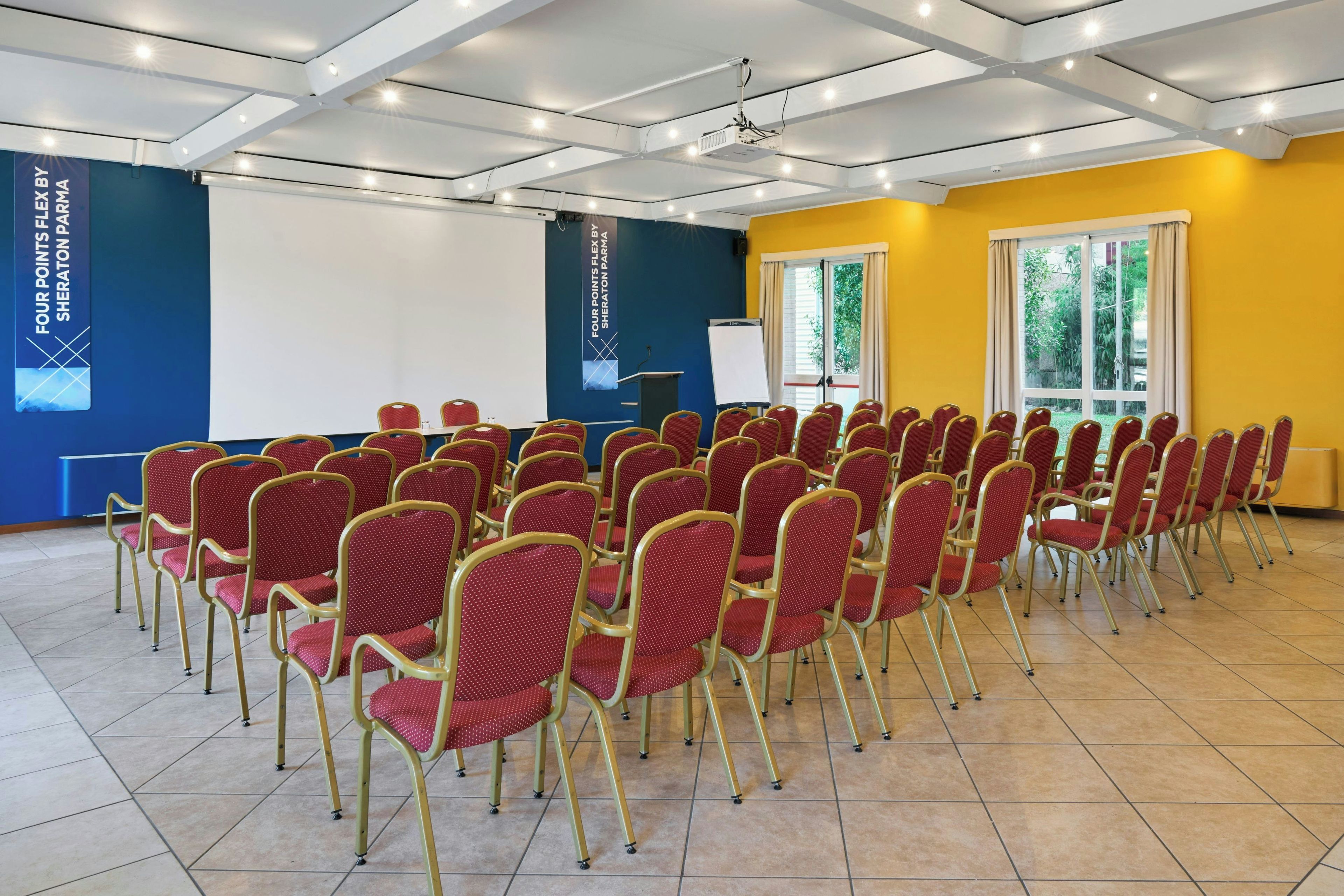 Une salle de conférence avec des rangées de chaises rouges, un projecteur et un tableau blanc.