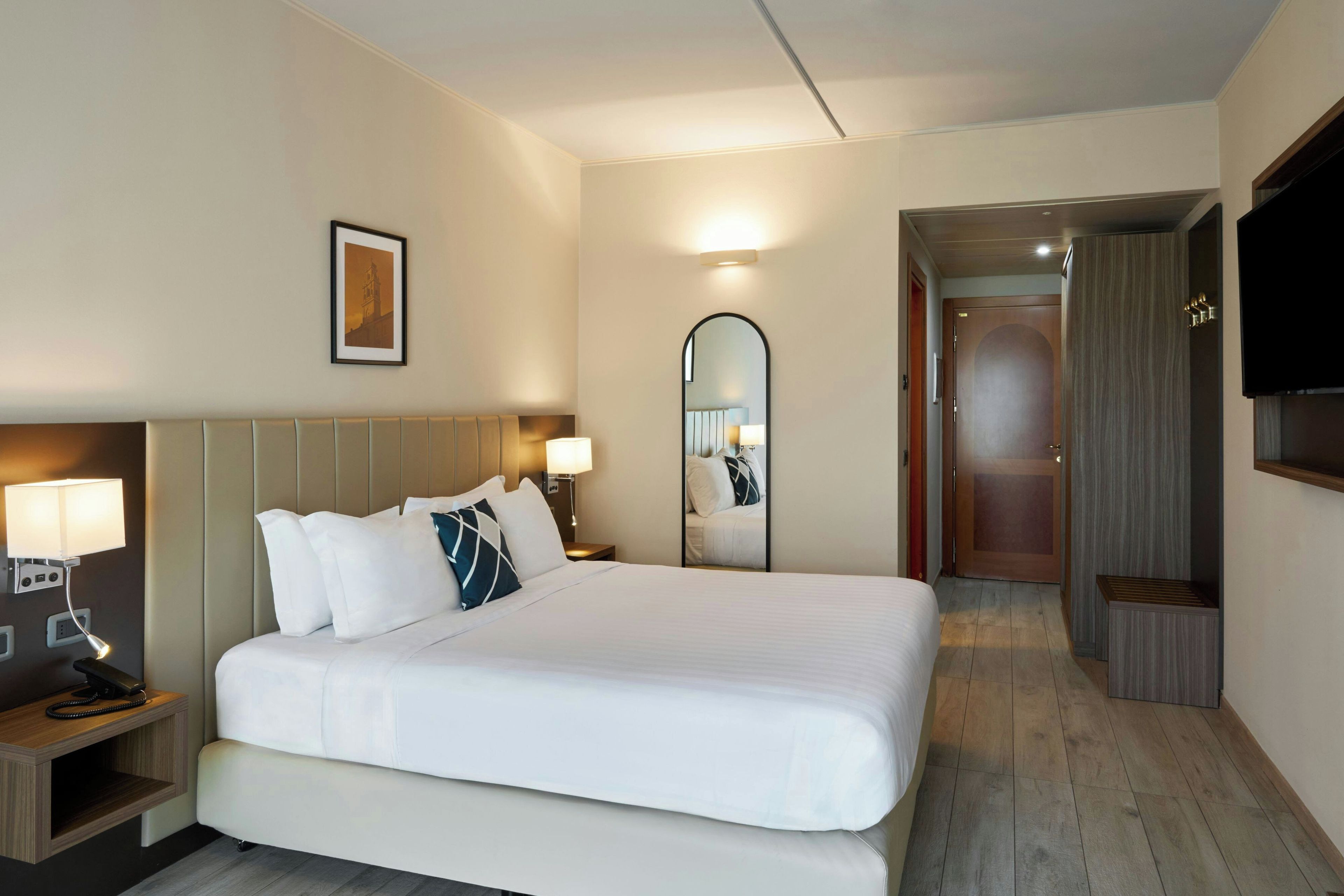 Una habitación de hotel con una cama grande, lámparas de noche, un espejo, un televisor y un cuadro en la pared.