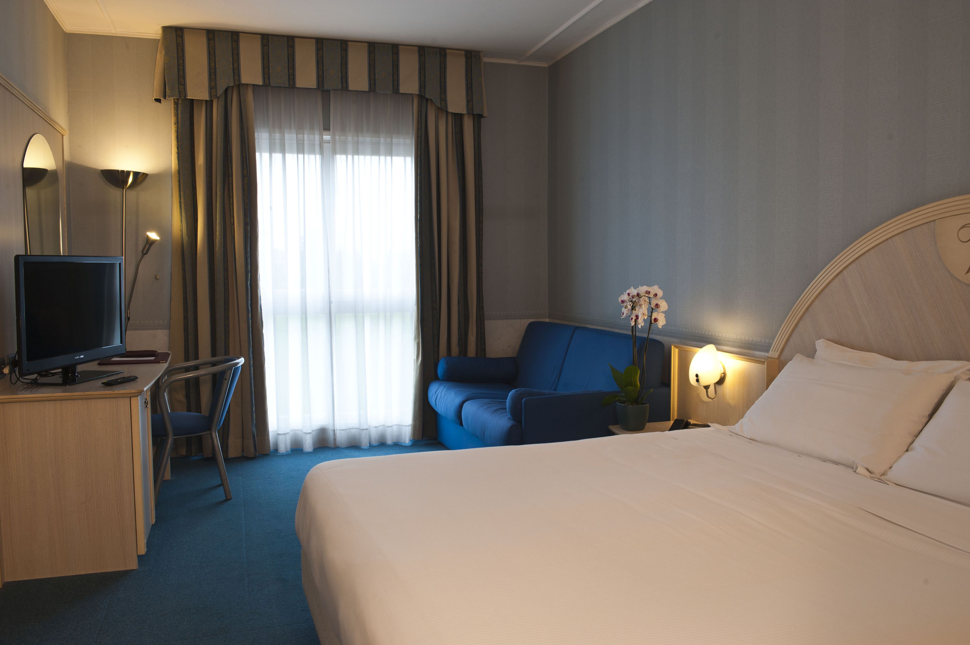Une chambre d’hôtel comprenant un lit, un bureau avec un ordinateur, une chaise, une télévision, une lampe et une fenêtre avec des rideaux.