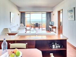 A hotel room with a balcony, a sofa, a dining table, a TV, and a minibar.