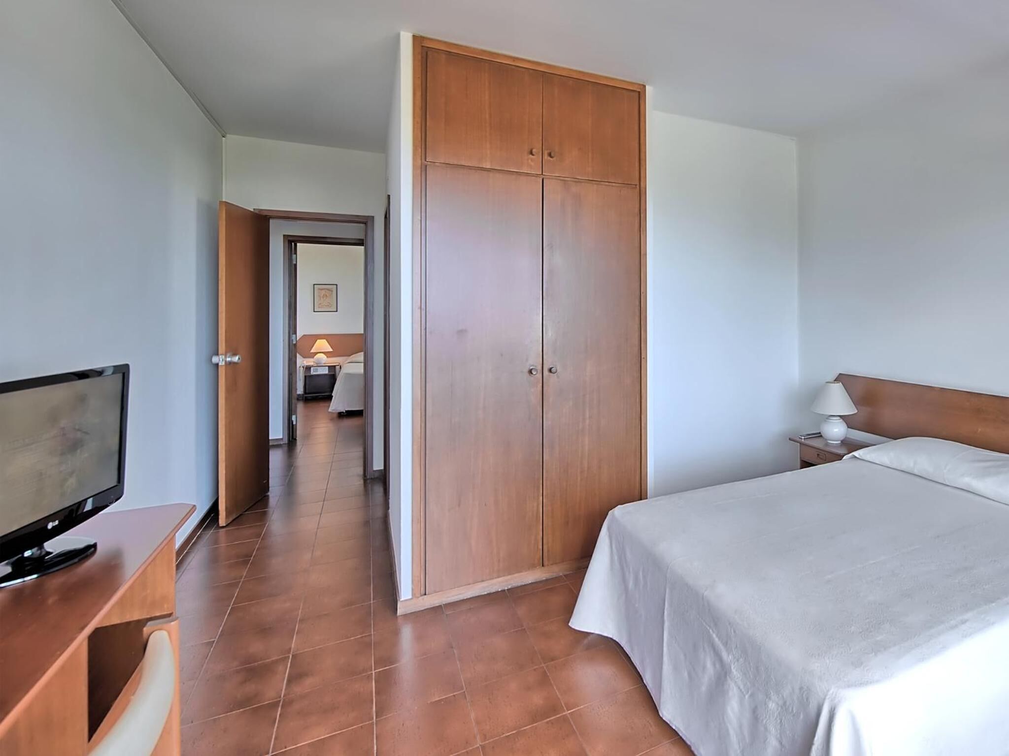 A hotel room with a bed, a desk with a TV, a wardrobe, and a nightstand with a lamp.