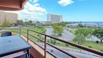 A balcony with a table and chairs, overlooking a parking area, a hotel, and a view of the sea.