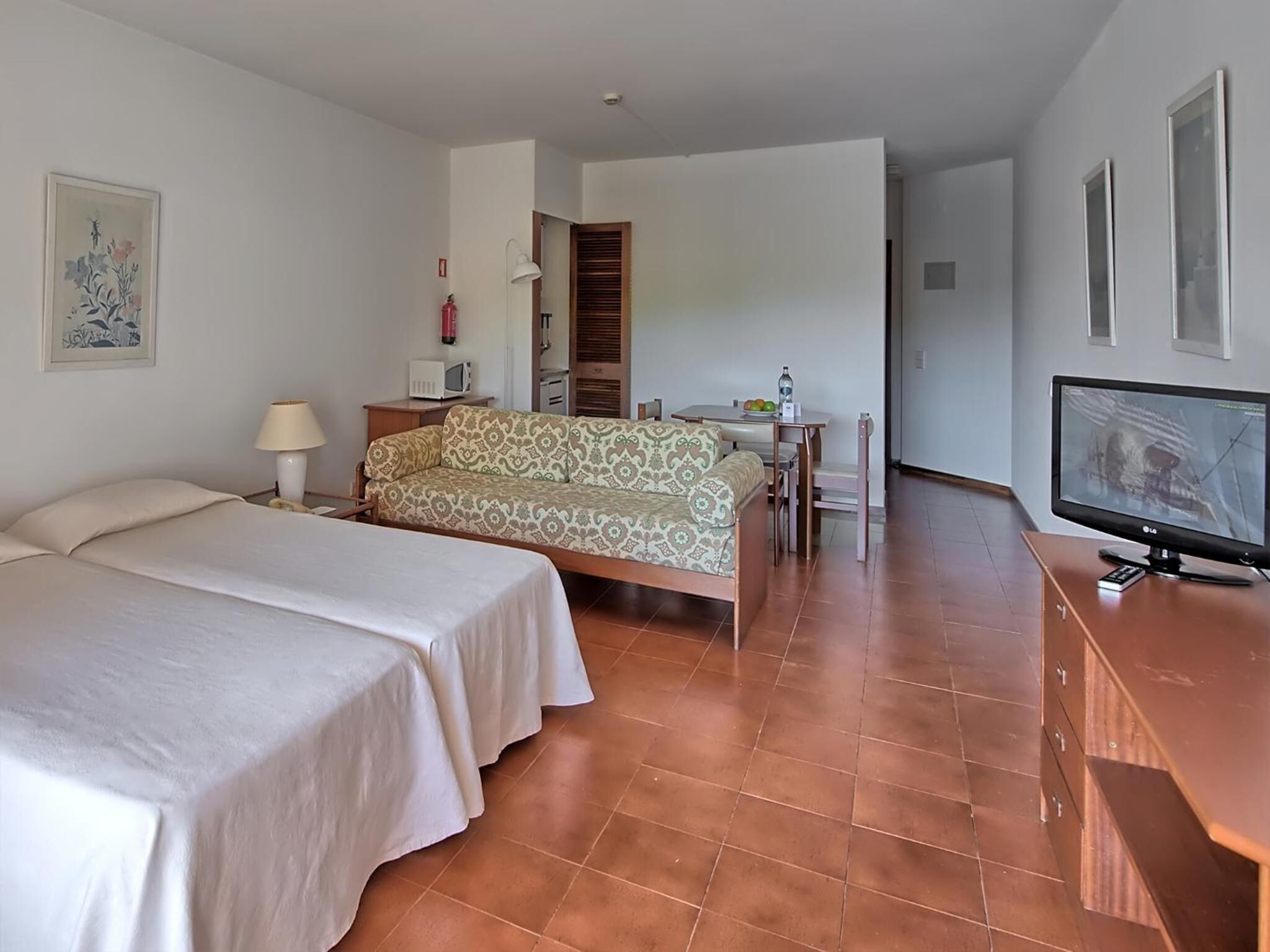 Habitación de hotel con dos camas, un sofá, un televisor, una mesita con lámpara y un cuadro en la pared.