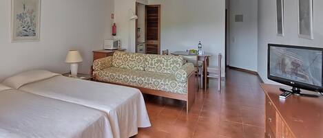Habitación de hotel con dos camas, un sofá, un televisor, una mesita con lámpara y un cuadro en la pared.