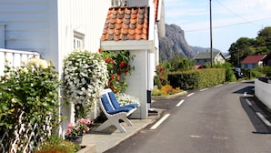 Une rue étroite avec un bâtiment blanc, une chaise bleue à l’extérieur, et une montagne en arrière-plan.