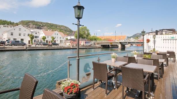 Une terrasse au bord de la rivière, aménagée avec des meubles en osier et des plantes en pot, d’où l’on peut admirer un pont et des bâtiments.