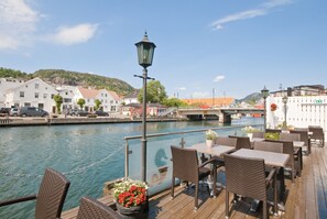 A riverside deck with wicker furniture and potted plants, overlooking a bridge and buildings.