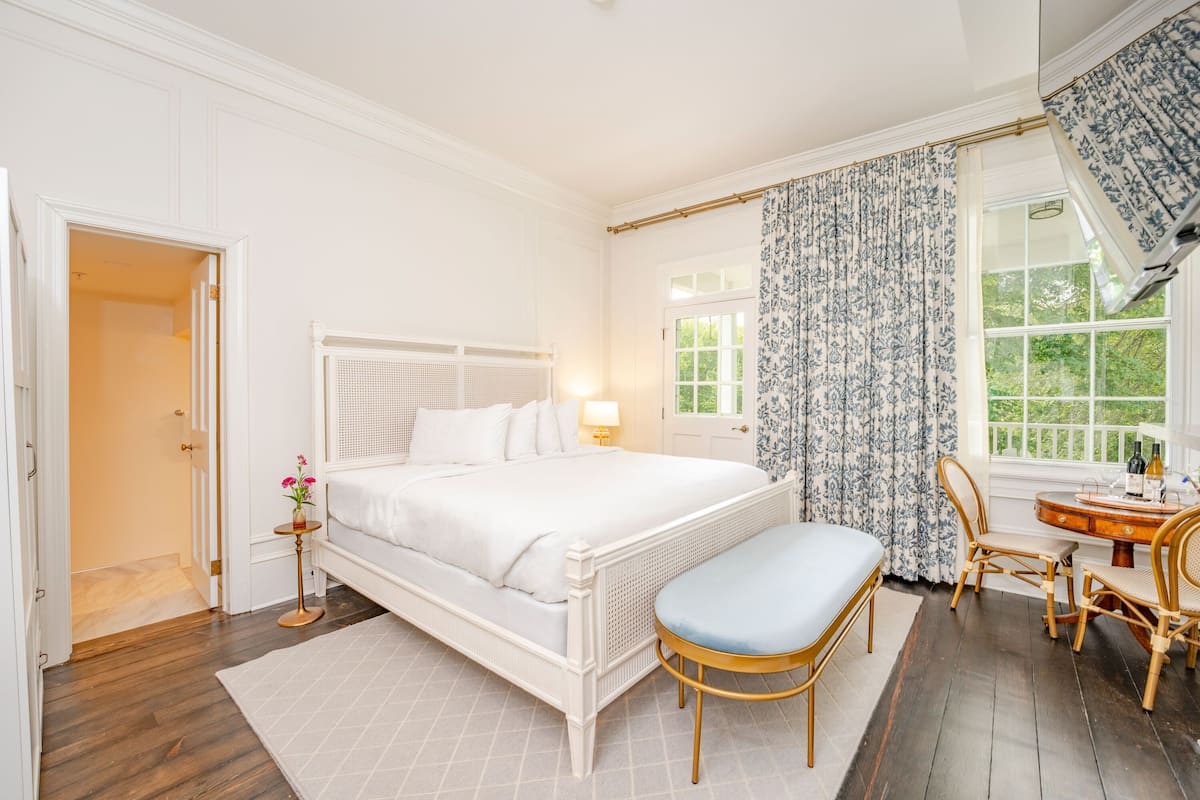 Un dormitorio con una cama blanca, una mesita de noche con lámpara, una silla y un ventanal con cortinas.