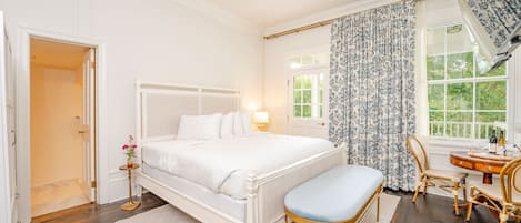 A bedroom with a white bed, a bedside table with a lamp, a chair, and a window with curtains.