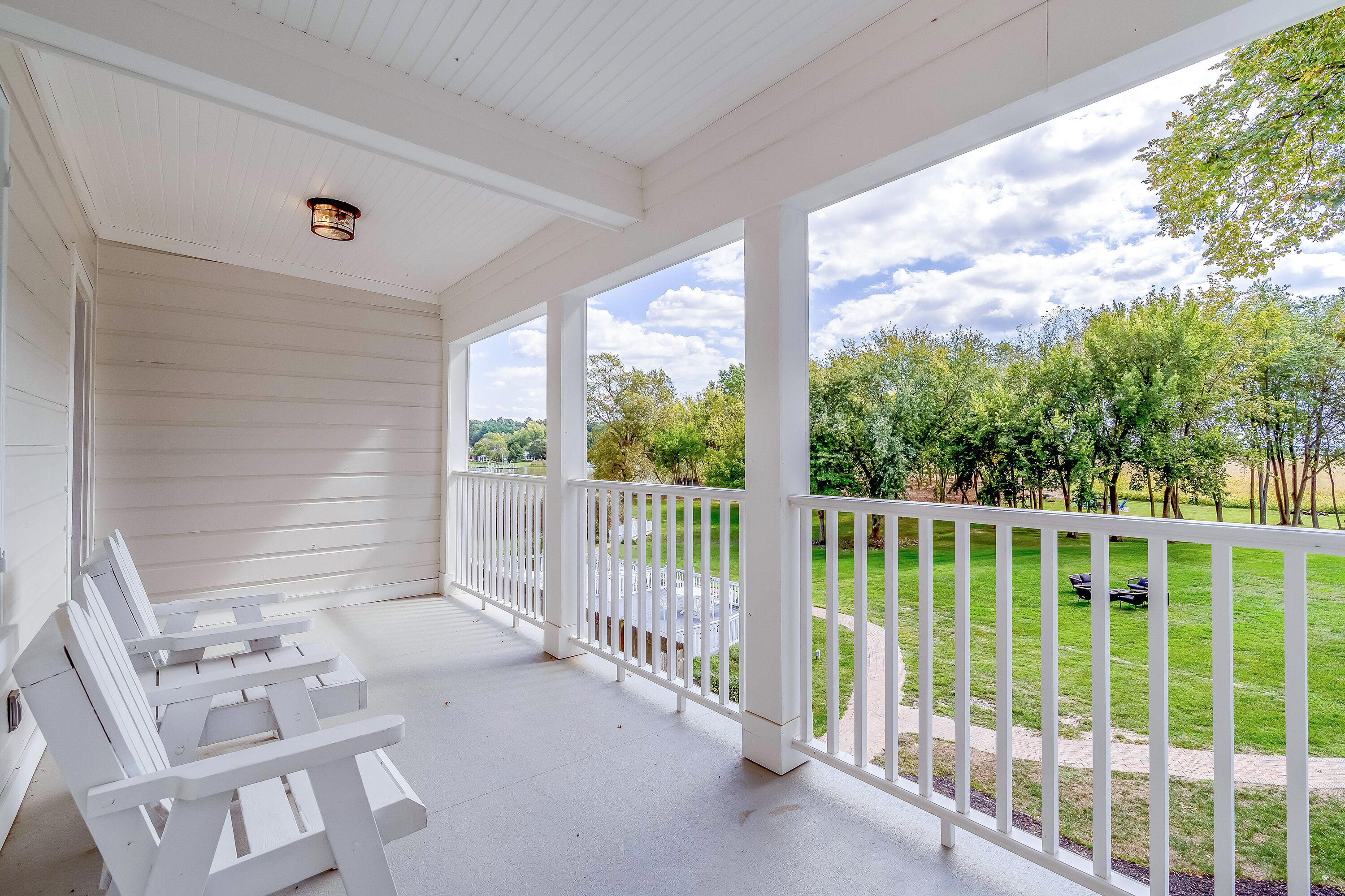 A covered porch with white railings and benches, overlooking a well-maintained lawn and trees.