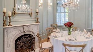 Una sala da pranzo con un tavolo rotondo apparecchiato per sei persone, circondato da sedie in legno. Si nota un camino con decorazioni elaborate, sopra il quale è appeso un grande specchio.