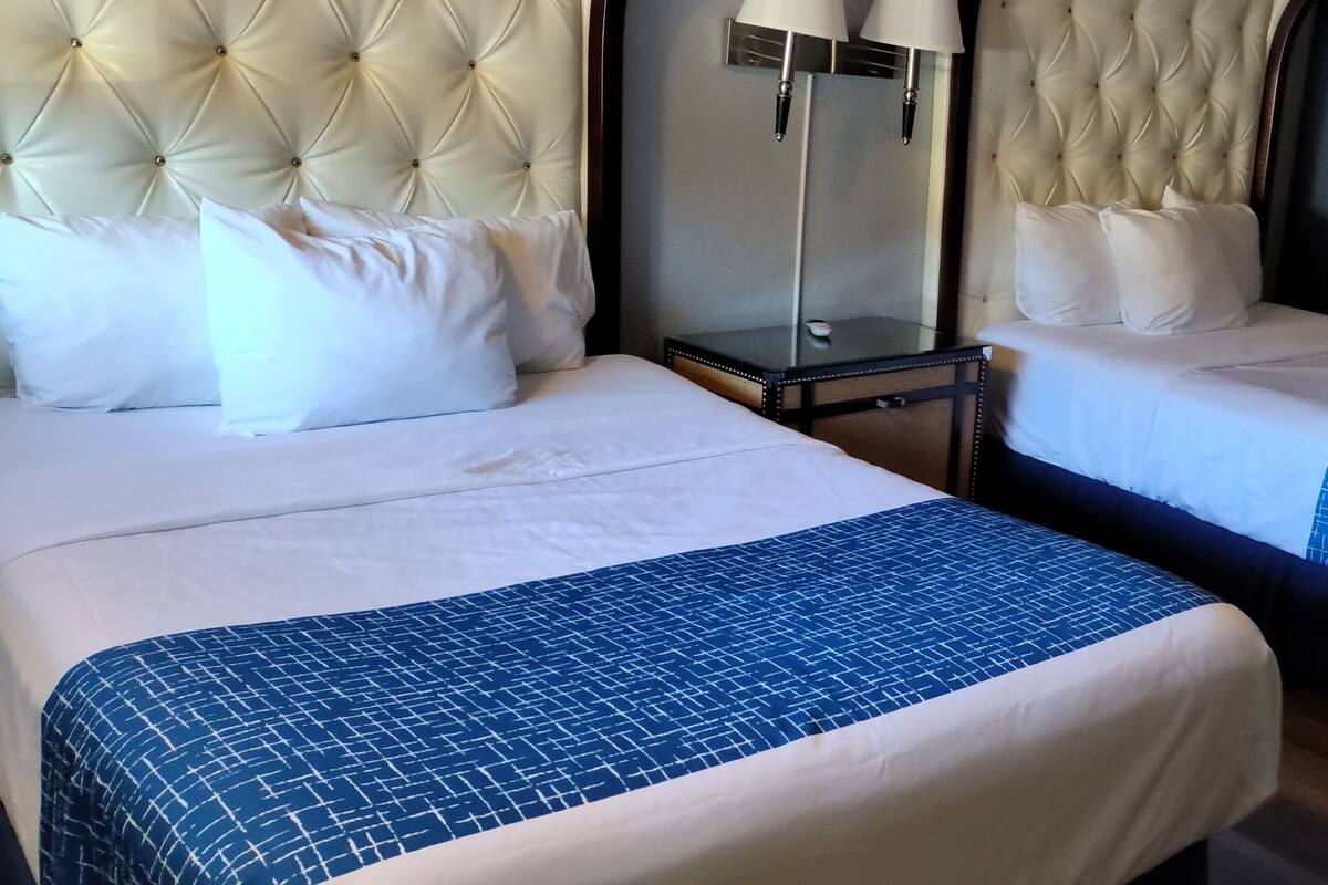 Deux lits avec du linge de lit blanc et des couvre-lits bleus, une tête de lit capitonnée, une table de chevet avec des lampes et un tableau encadré au mur.