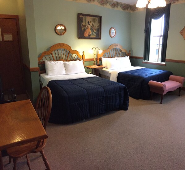 A room with two beds, a wooden table, a chair, a small table with a lamp, and a window with curtains.