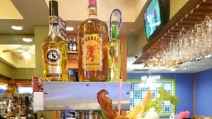 Ambiente de bar com um cocktail Bloody Mary, garrafas de bebidas alcoólicas e uma tela de televisão ao fundo.