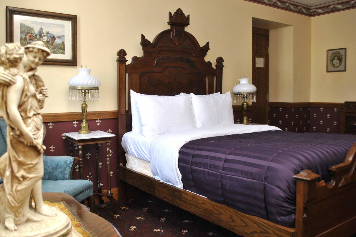 Ein Schlafzimmer mit einem hölzernen Bett, weißer Bettwäsche, einer violetten Decke, zwei Nachttischlampen, einem blauen Sessel und einer Statue.