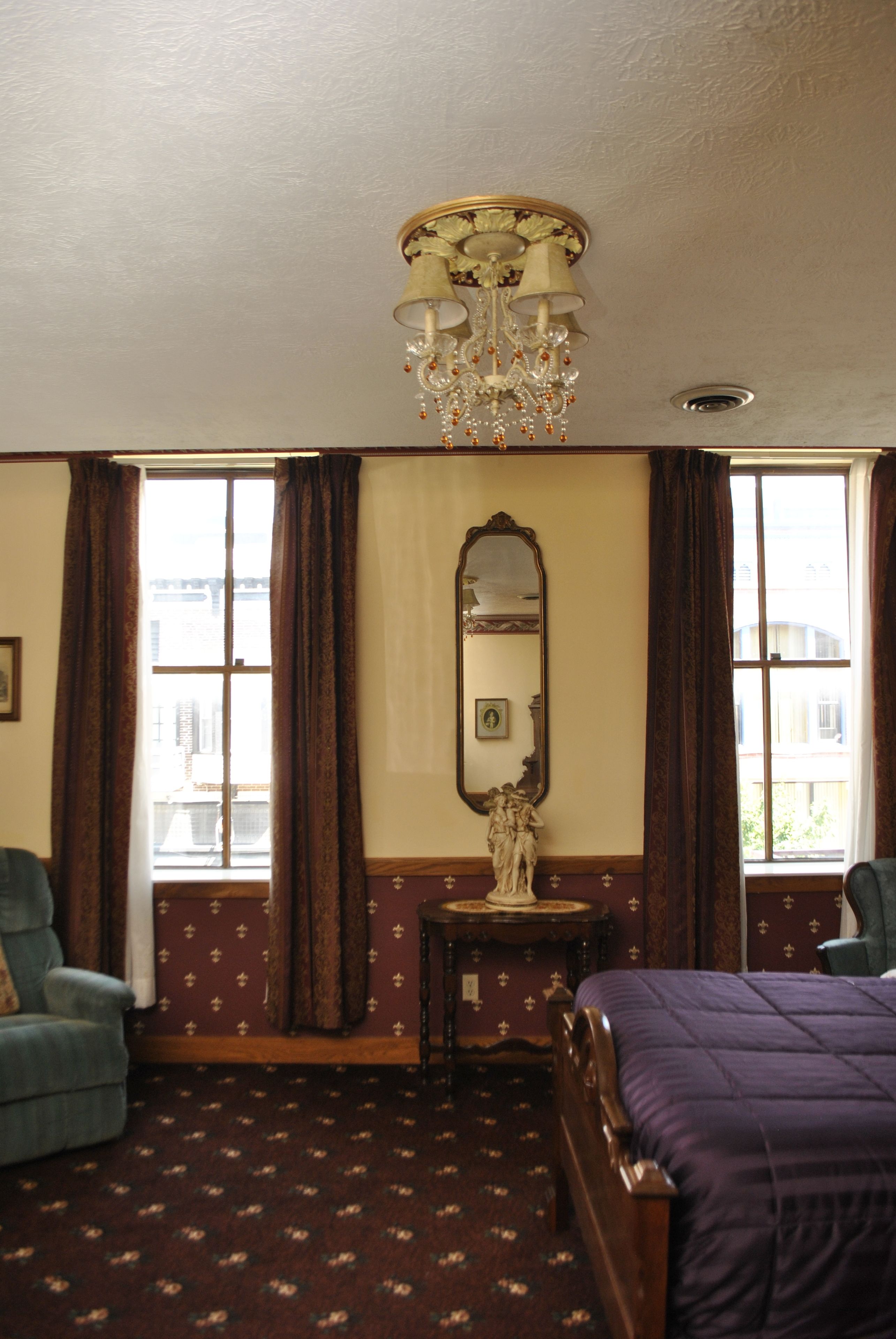 A hotel room with a large window, a chandelier, a mirror, a wooden bed, and a blue armchair.