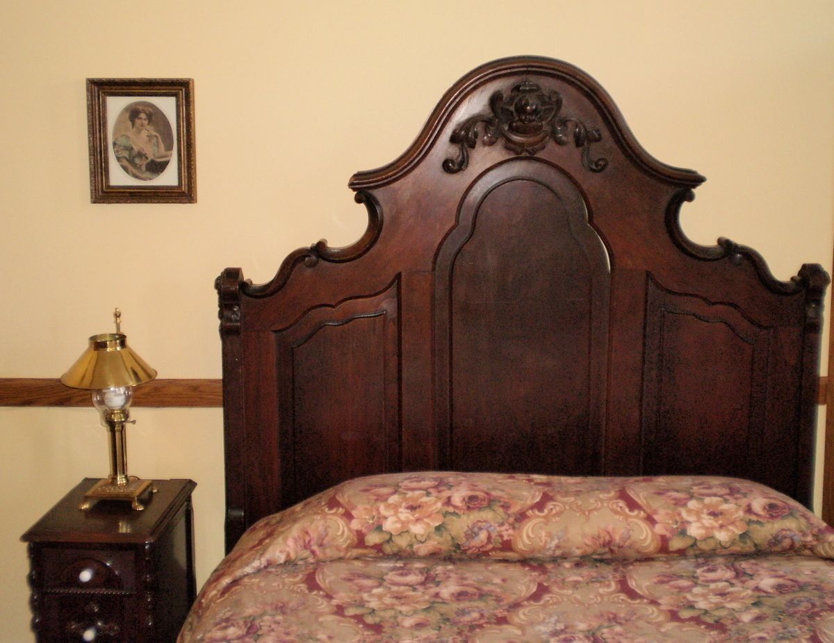 Une chambre à coucher avec une tête de lit en bois, une couvre-lit à motifs floraux, une table de chevet avec une lampe et un tableau encadré accroché au mur.