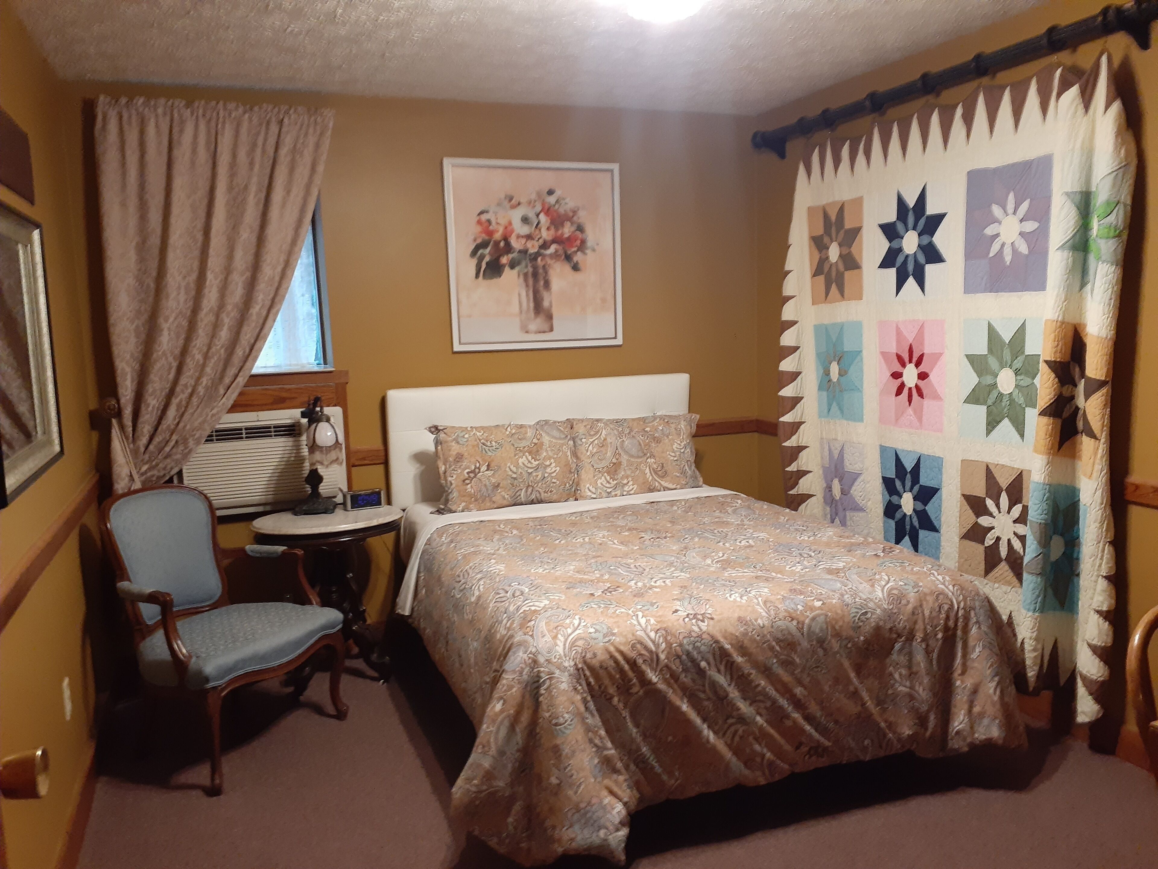 A bedroom with a bed, a chair, a bedside table, a window with curtains, a wall-mounted air conditioner, and a floral painting.