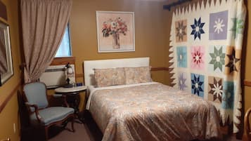 A bedroom with a bed, a chair, a bedside table, a window with curtains, a wall-mounted air conditioner, and a floral painting.