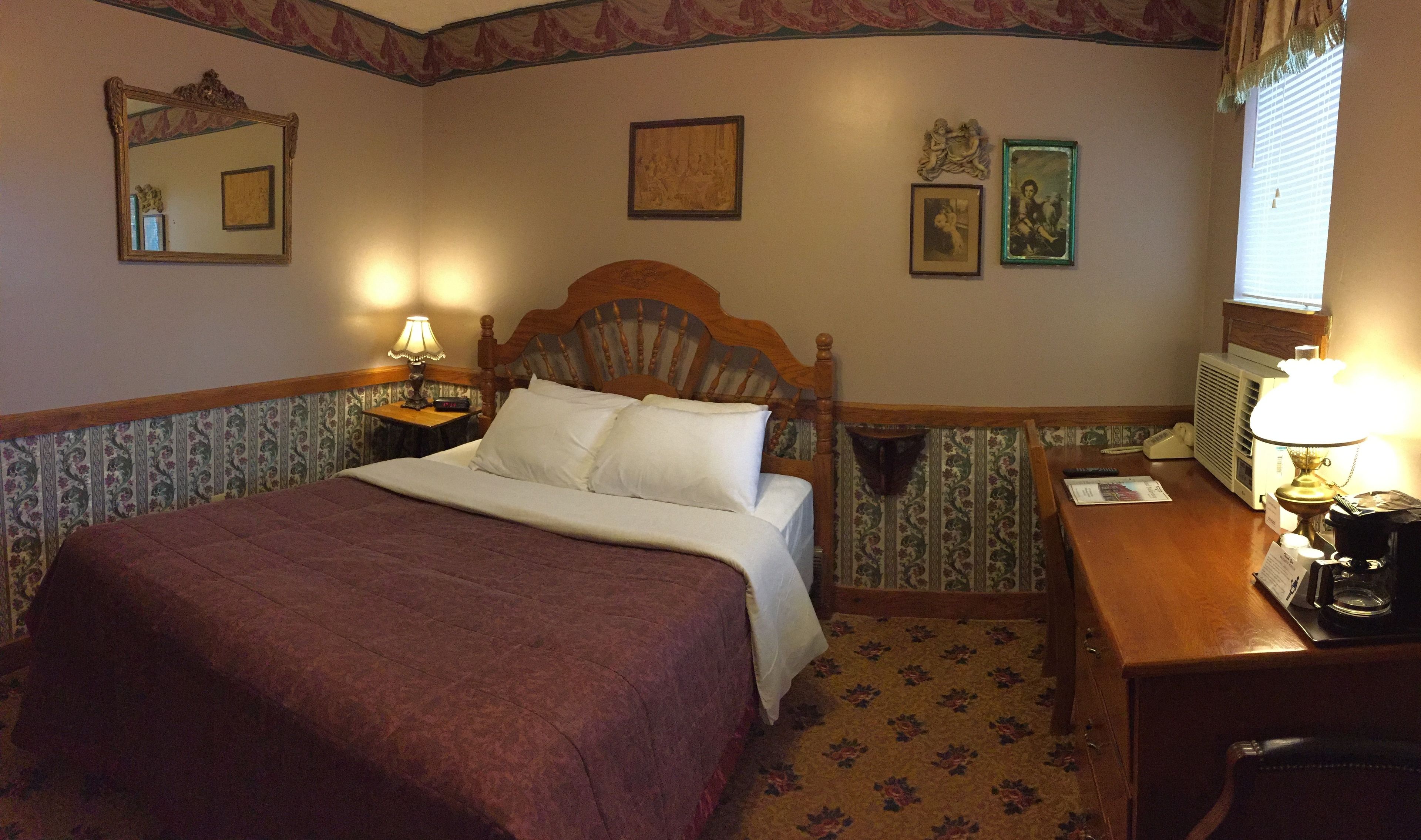 Quarto de hotel com uma cama grande, duas luminárias laterais, uma cabeceira de madeira, uma mesa pequena com uma luminária, um espelho e uma janela com persianas.