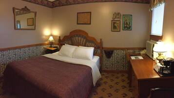 Quarto de hotel com uma cama grande, duas luminárias laterais, uma cabeceira de madeira, uma mesa pequena com uma luminária, um espelho e uma janela com persianas.