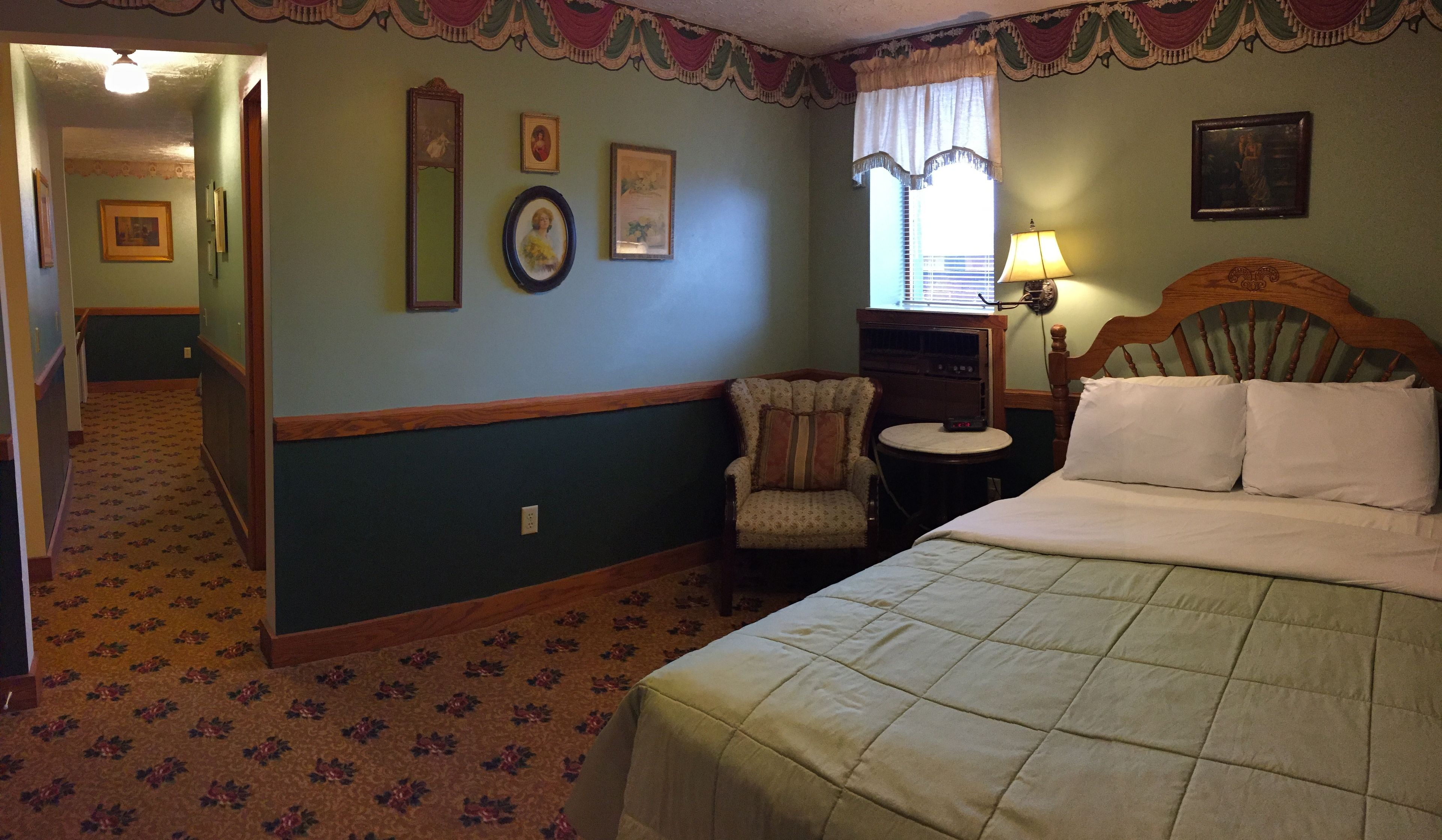 A bedroom with a bed, a chair, a desk with a lamp, and a window with curtains.